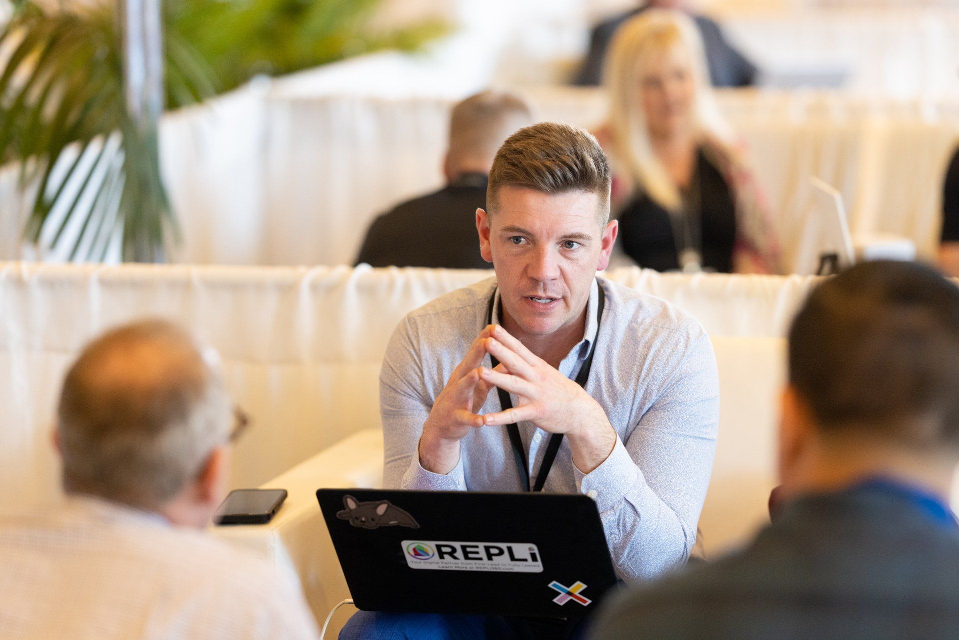 A man is sitting at a table with a laptop and talking to another man.