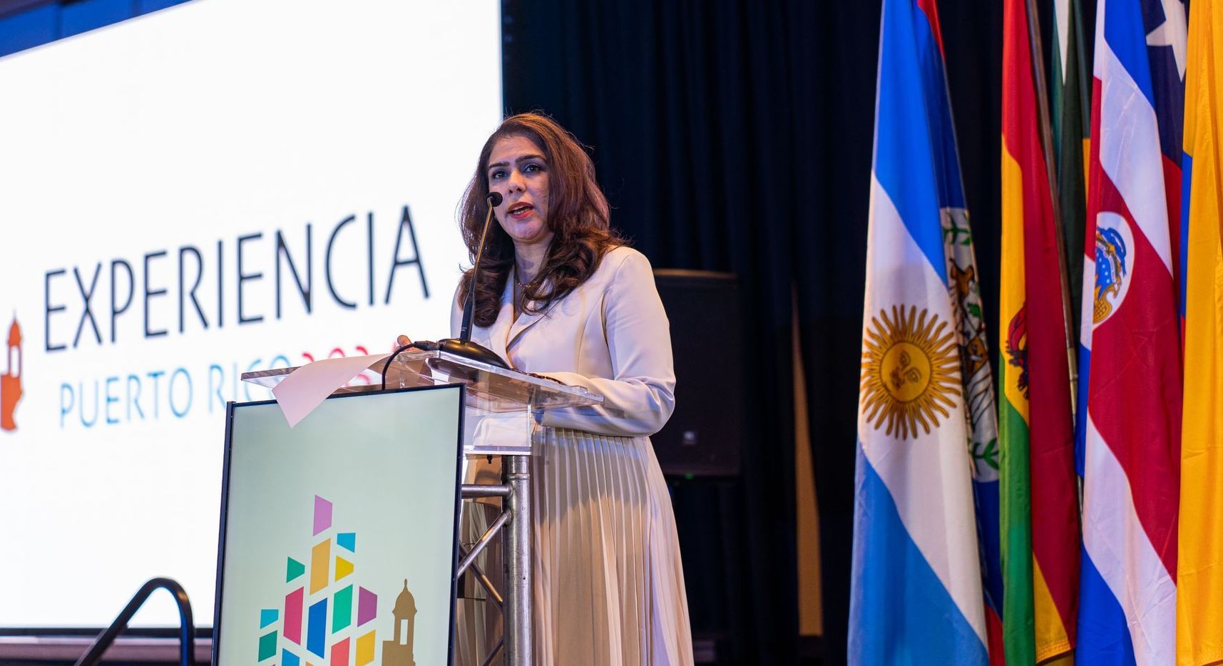 A woman is standing at a podium giving a speech in front of flags.